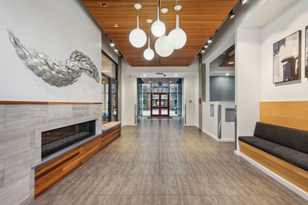 A modern building lobby featuring a fireplace with metal wall art, a central corridor with globe pendant lights, and built-in benches.