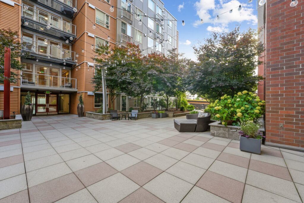 A paved apartment courtyard featuring lounge chairs, potted plants, and overhead string lights nestled between modern wooden and brick buildings.