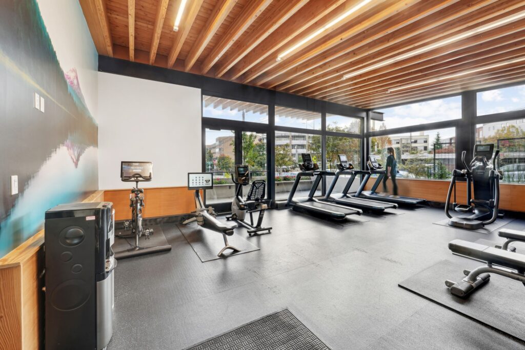 A gym with a stationary bike, a rowing machine, three treadmills, and a weight bench, overlooking a street through large windows.