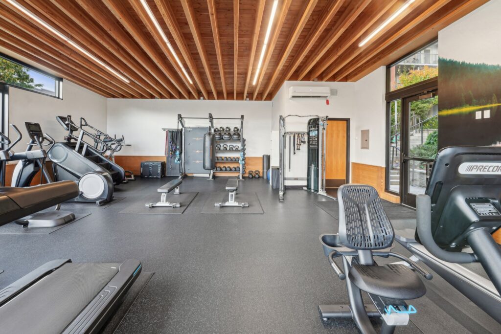 A spacious home gym with treadmills, a recumbent bike, weight benches, a punching bag, and a rack of kettlebells under a wooden-beamed ceiling.