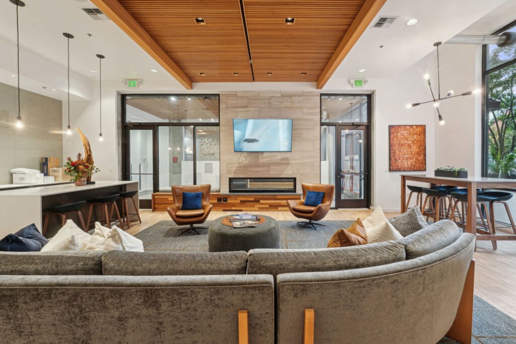 A modern lounge featuring a gray sectional sofa, two leather swivel chairs facing a wall-mounted television and fireplace, and a kitchen island with bar stools.