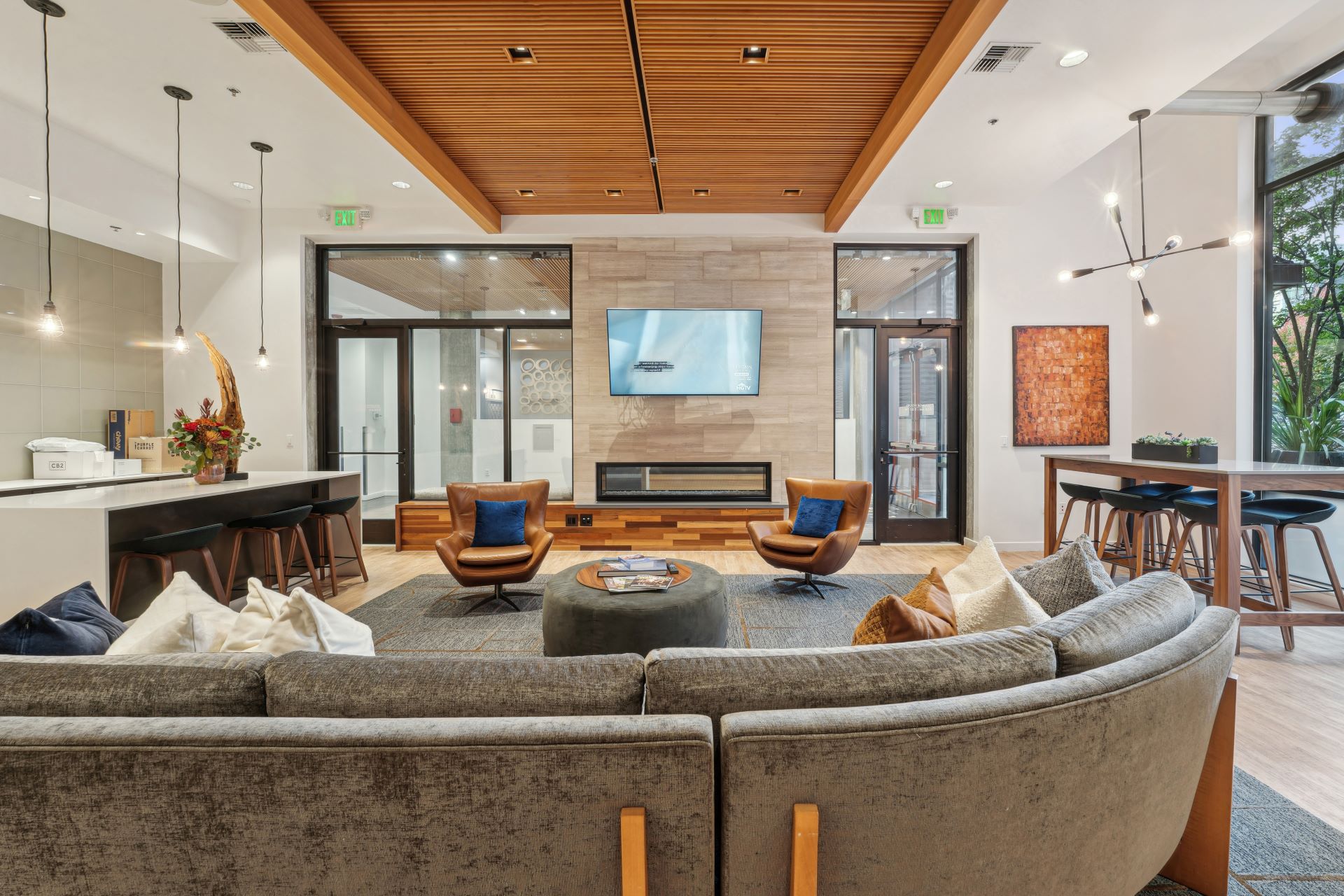 A modern lounge featuring a gray sectional sofa, two leather swivel chairs facing a wall-mounted television and fireplace, and a kitchen island with bar stools.