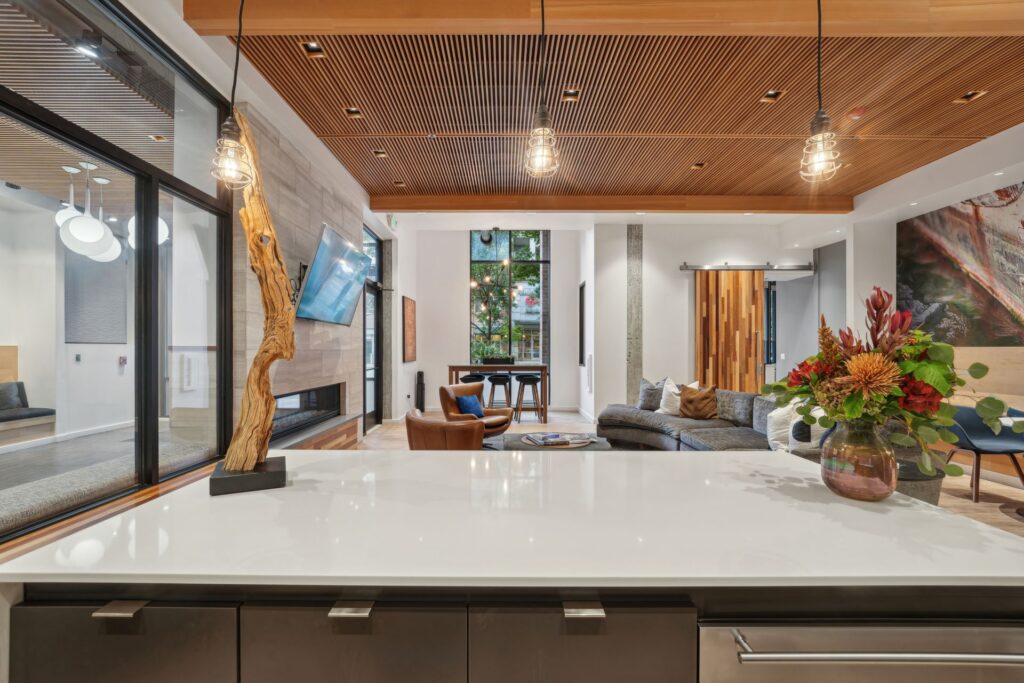 A view from a kitchen counter looking into a modern, open-concept living room with a slatted wood ceiling, a flat-screen television above a fireplace, and a large sectional sofa.
