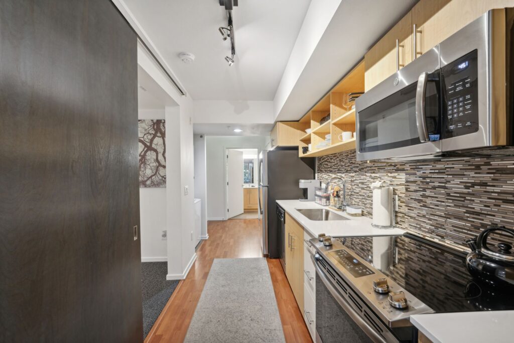 A galley-style kitchen with wood cabinets, stainless steel appliances, and a tiled backsplash, looking through to a hallway and a laundry area.