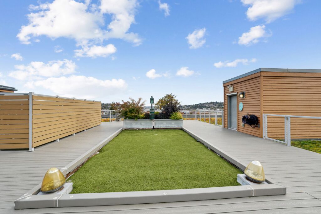 A rooftop deck featuring a central rectangular patch of artificial turf bordered by a wooden walkway and a small service building.