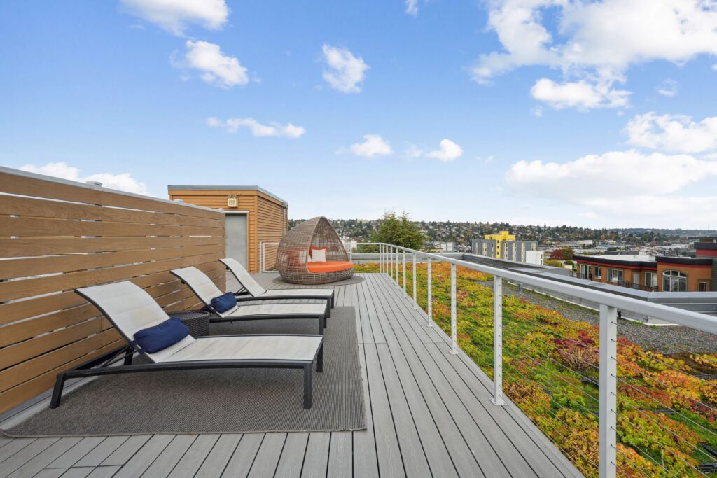 Three lounge chairs on a grey wooden rooftop deck next to a circular wicker lounge seat, overlooking a green roof and city skyline.