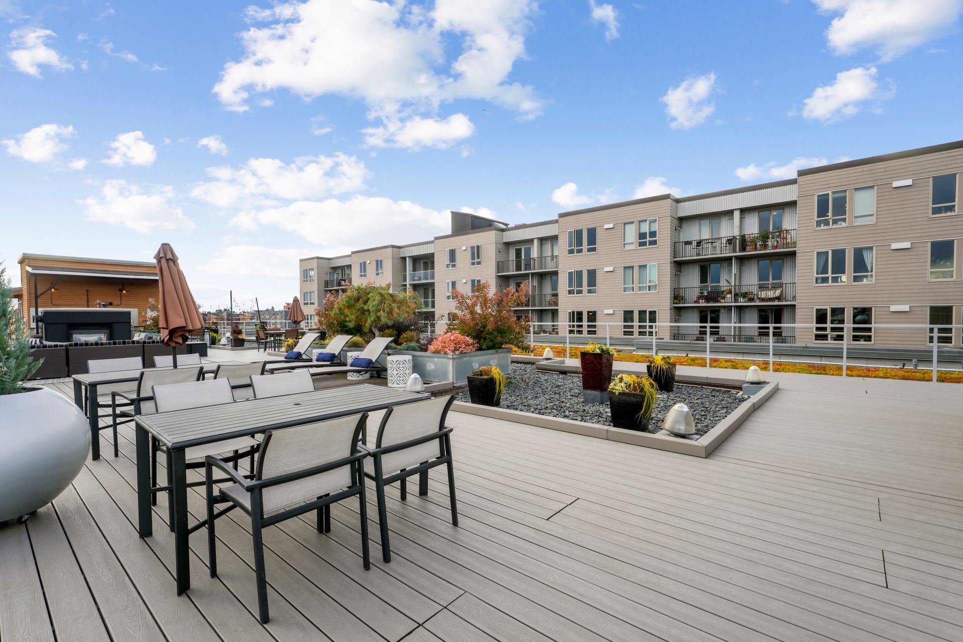 Rooftop terrace with dining tables, lounge chairs, and planters against an apartment building.