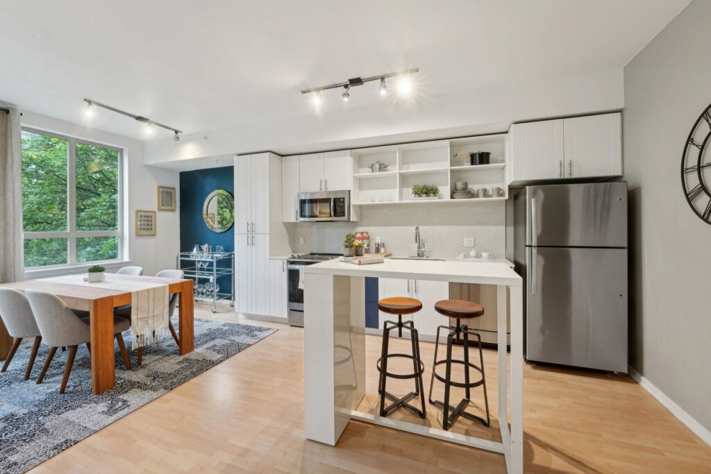 Open-concept kitchen and dining area featuring a white kitchen island with two stools, stainless steel appliances, white cabinetry, and a wooden dining table for four near a large window.