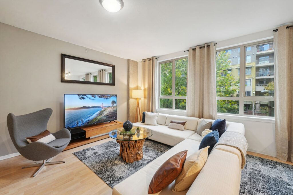 A living room featuring a white sectional sofa, a glass-top coffee table with a wood base, a grey armchair, and a television mounted above a media console.