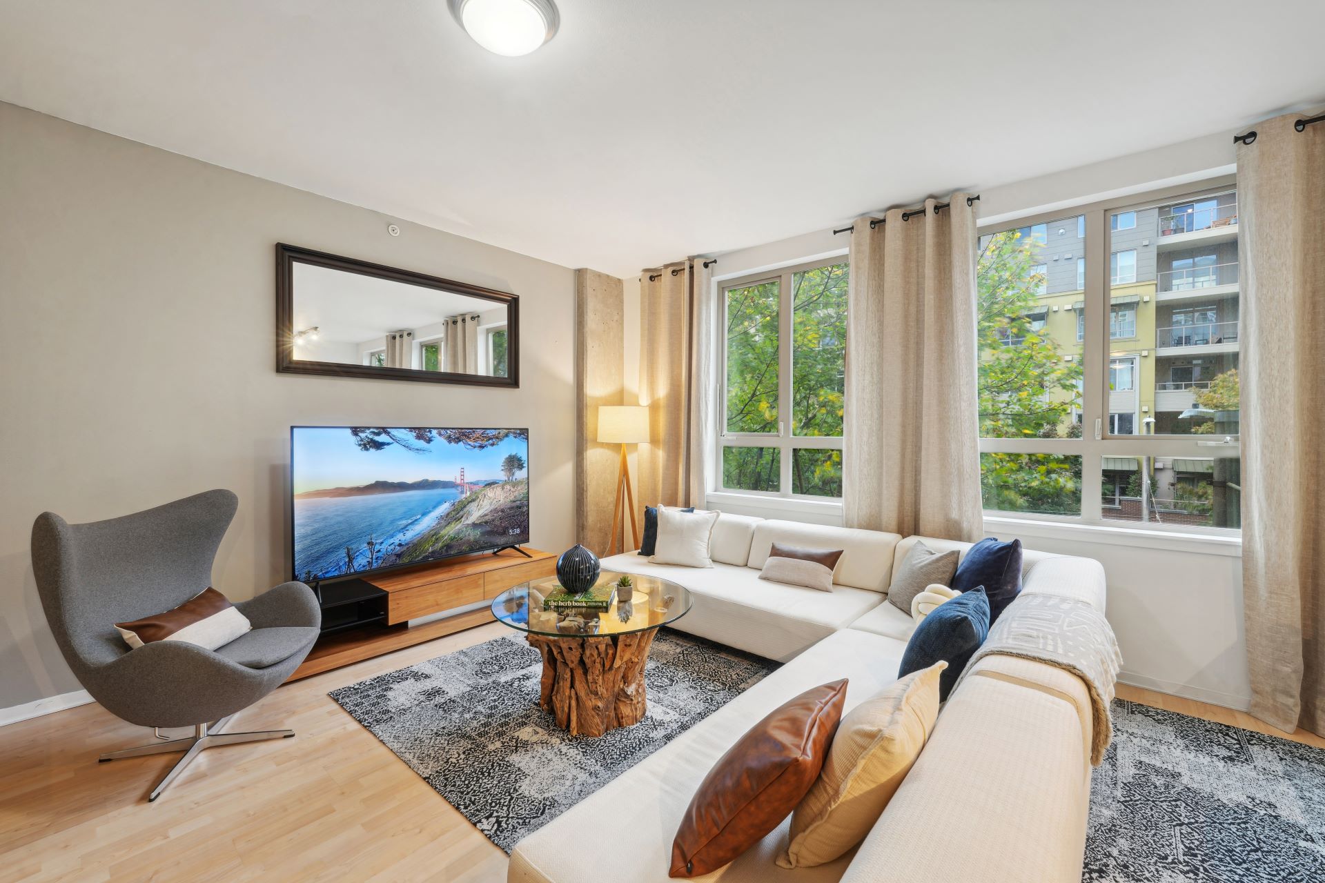 A living room featuring a white sectional sofa, a glass-top coffee table with a wood base, a grey armchair, and a television mounted above a media console.