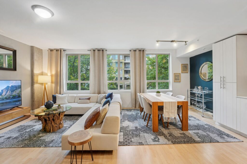 An open-concept living and dining area featuring a sectional sofa, a coffee table with a tree-stump base, a wooden dining table with six chairs, and a bar cart against a dark blue accent wall.