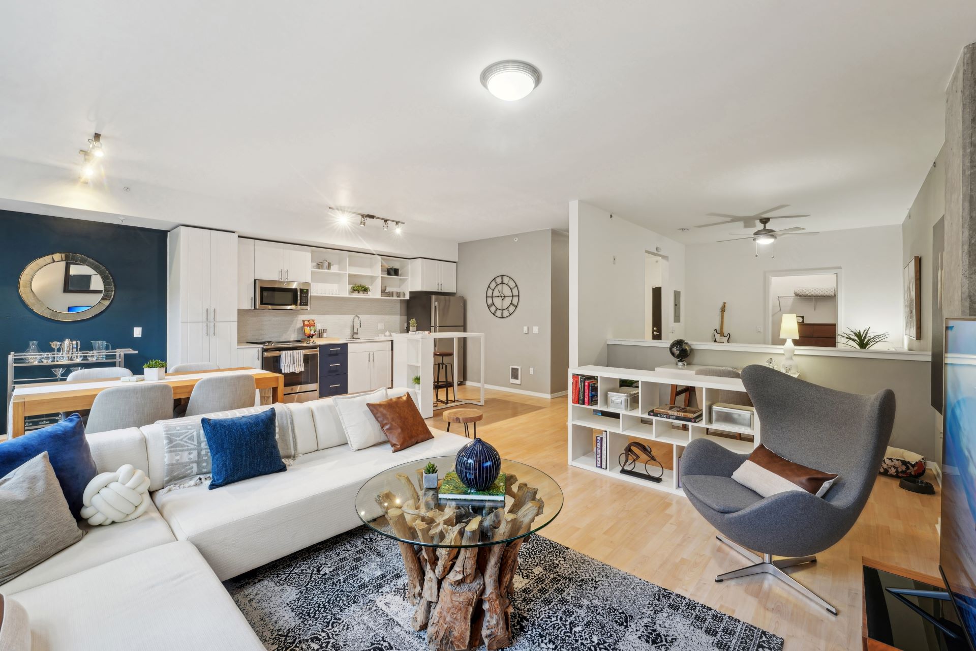 Open-concept living area with a white sectional sofa, a glass-top coffee table with a driftwood base, and an adjacent kitchen with white cabinetry.