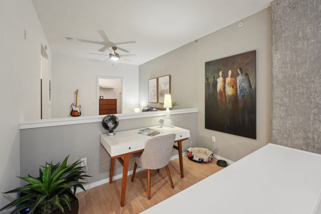 A white desk with a globe and lamp sits against a partial wall in an apartment living area, featuring a large abstract painting of figures on the adjacent wall.