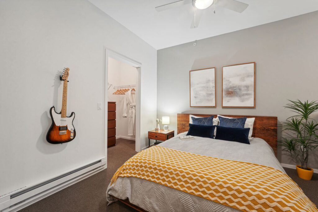 A bedroom featuring a bed with a wooden headboard, yellow patterned throw, and two abstract art pieces above it, with an electric guitar mounted on the wall and an open walk-in closet nearby.