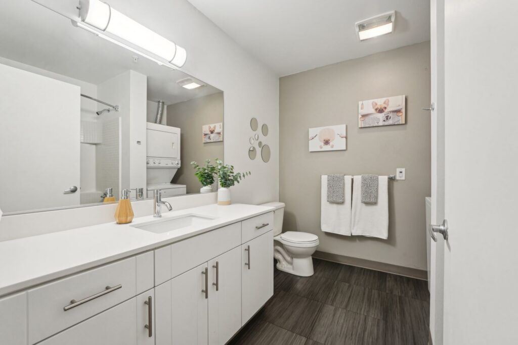 A bathroom featuring a white vanity with a single sink, a toilet, a washer and dryer unit, and two dog-themed wall canvases.
