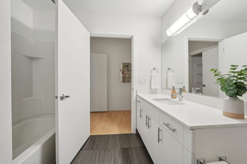 A modern bathroom featuring a white vanity with silver pulls, a rectangular mirror, a bathtub, and a doorway leading to a room with light wood flooring.