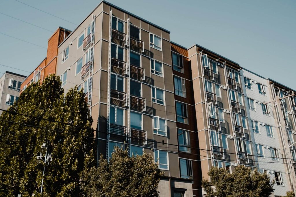 A modern multi-story apartment building with large windows and balconies, partially obscured by trees in the foreground.