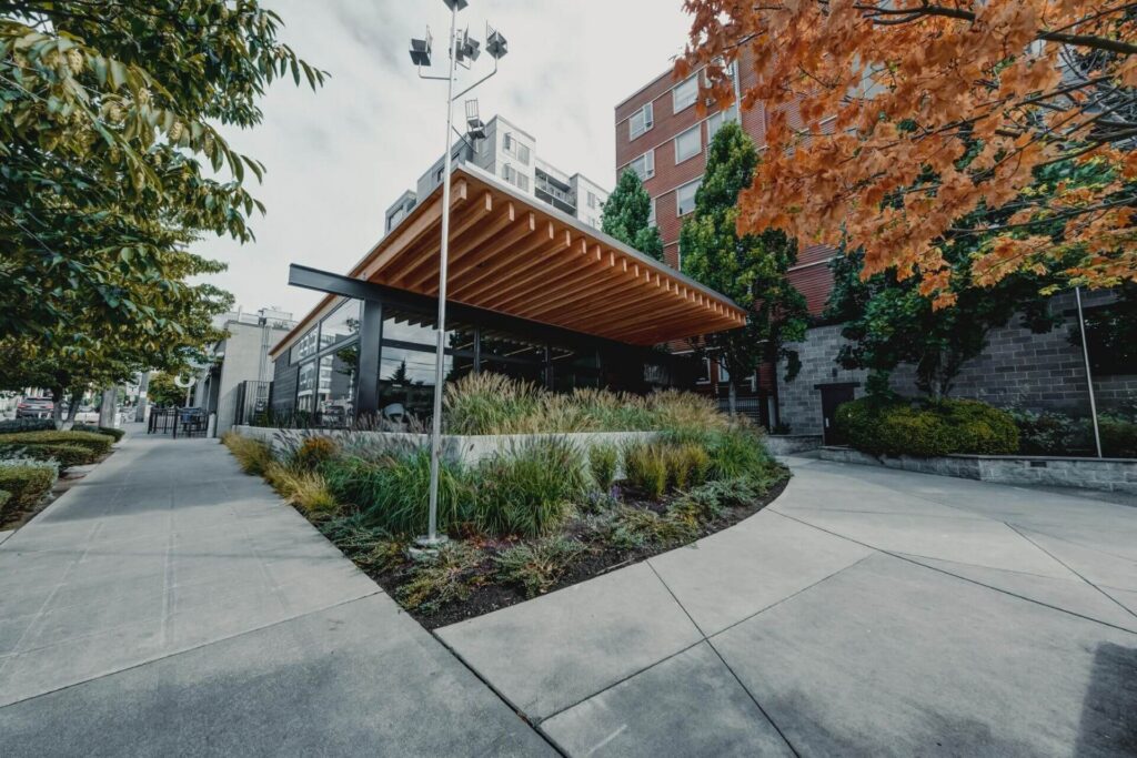 A modern building with a large wooden sloped roof and glass walls, surrounded by landscaping and concrete walkways.
