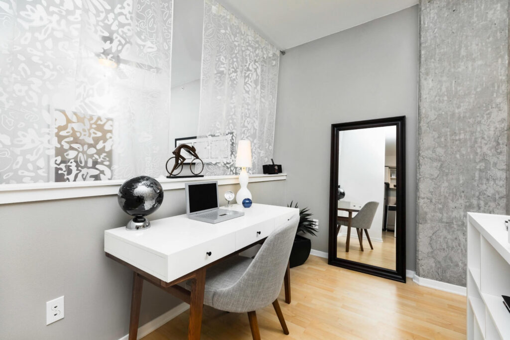 A white desk with a laptop, globe, and lamp, accompanied by a gray chair and a full-length floor mirror in a room with a concrete pillar.