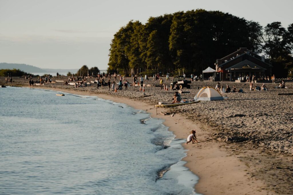 People relax on a sandy beach near a treeline and a small building, with a child playing near the waters edge.